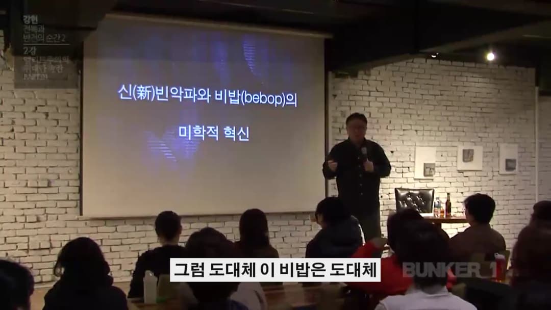 강연자가 스크린 앞에 서서 청중을 향해 이야기하고 있으며, 스크린에는 '신빈악파와 비밥의 미학적 혁신'이라는 문구가 적혀 있다