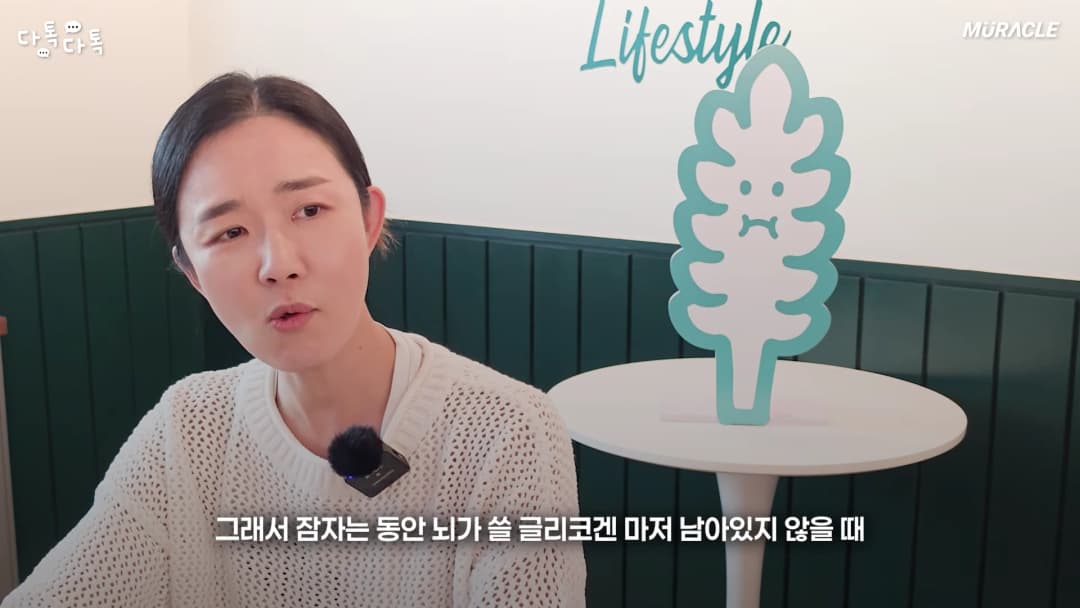 흰색 니트를 입은 여성이 인터뷰 중이며 옆에는 나뭇잎 모양의 캐릭터 조형물이 놓여 있다.