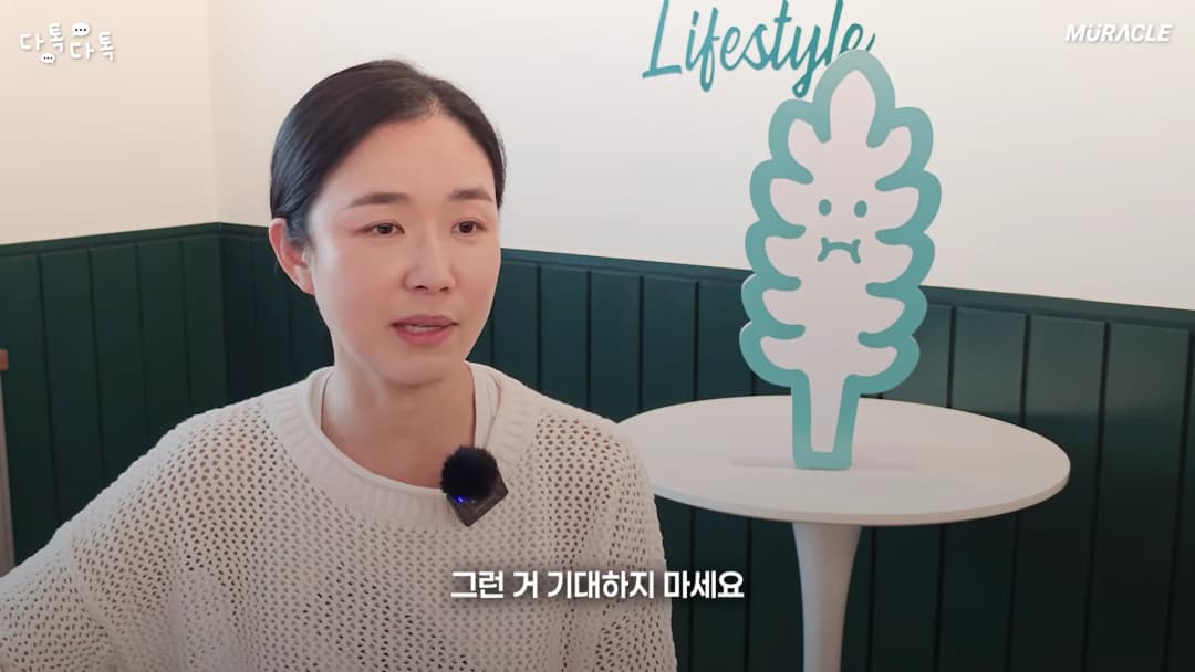 흰색 니트를 입은 여성이 인터뷰 중이며, 옆에는 나뭇잎 모양의 캐릭터 조형물이 놓여 있다.