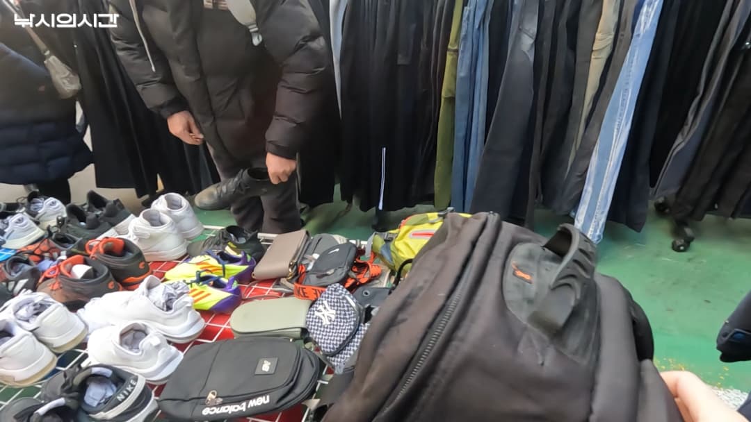 동묘 구제시장의 노점 매대에 다양한 중고 신발과 가방이 진열되어 있고, 한 사람이 물건을 살펴보고 있다.